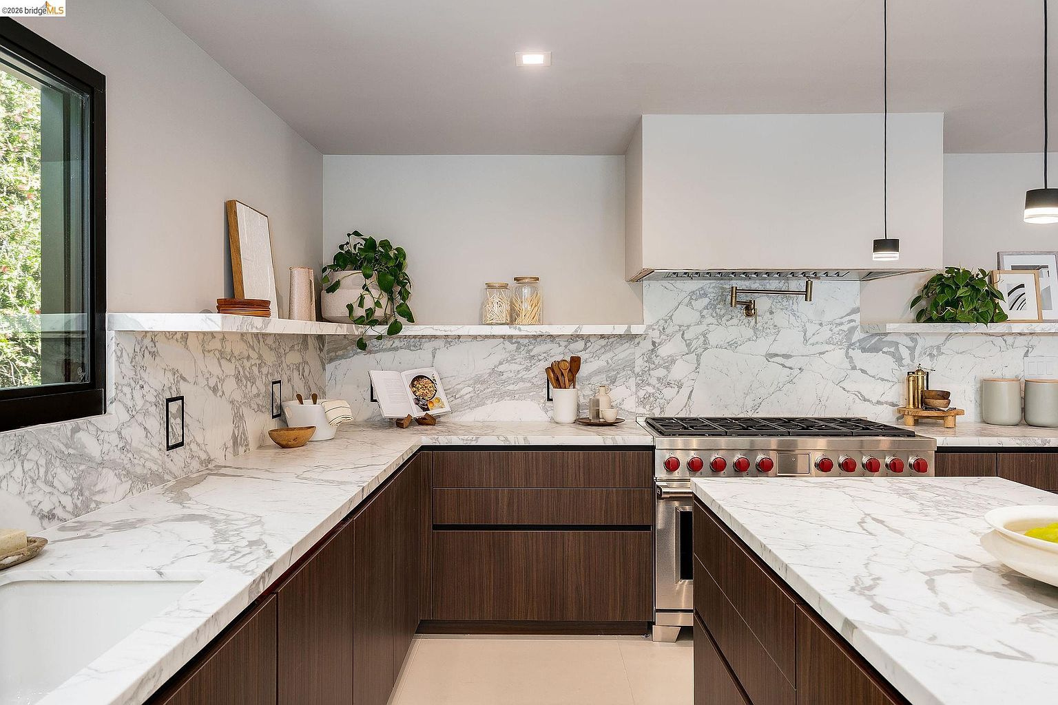 This sophisticated, modern kitchen features rich, dark wood cabinetry contrasted by striking white marble countertops and a matching full-height backsplash. A professional-grade range with red knobs serves as the focal point, complemented by minimalist floating shelves and sleek pendant lighting. The perspective is a wide, eye-level shot that emphasizes the clean lines, high-end finishes, and open, airy layout of this luxury culinary space.