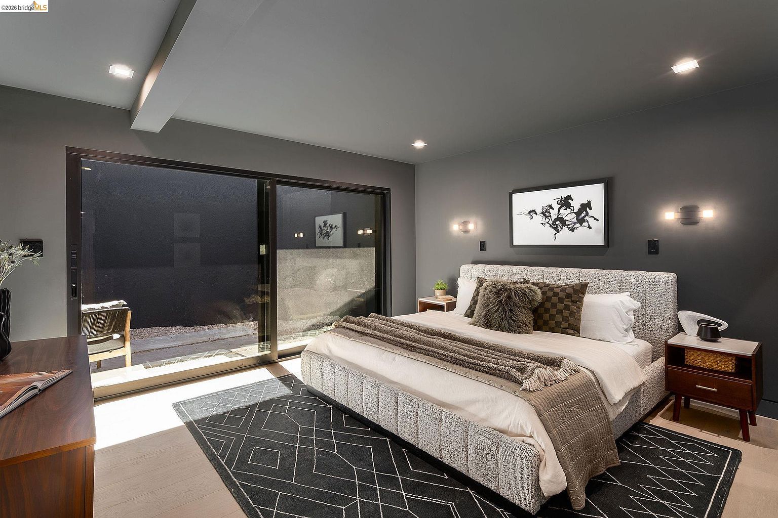 This sophisticated primary bedroom features a contemporary design with a dark, moody color palette and a large, textured upholstered bed as the focal point. A geometric patterned rug anchors the space, while floor-to-ceiling sliding glass doors provide a seamless transition to a private outdoor patio area. The room is accented by modern wall sconces and a minimalist art piece, creating a serene and luxurious atmosphere.
