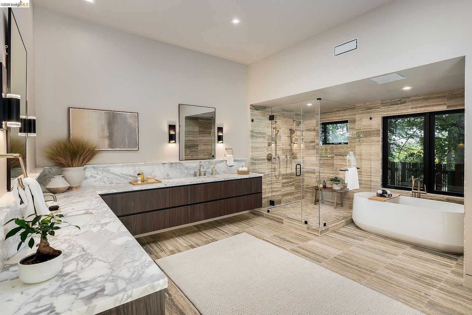 This luxurious primary bathroom features a sleek, modern design with a floating double vanity topped with white marble, complemented by dark wood cabinetry. A spacious walk-in glass shower with textured stone tile walls sits adjacent to a contemporary freestanding soaking tub, creating a spa-like atmosphere. The room is bathed in natural light from a large window, highlighting the elegant marble flooring and sophisticated lighting fixtures.