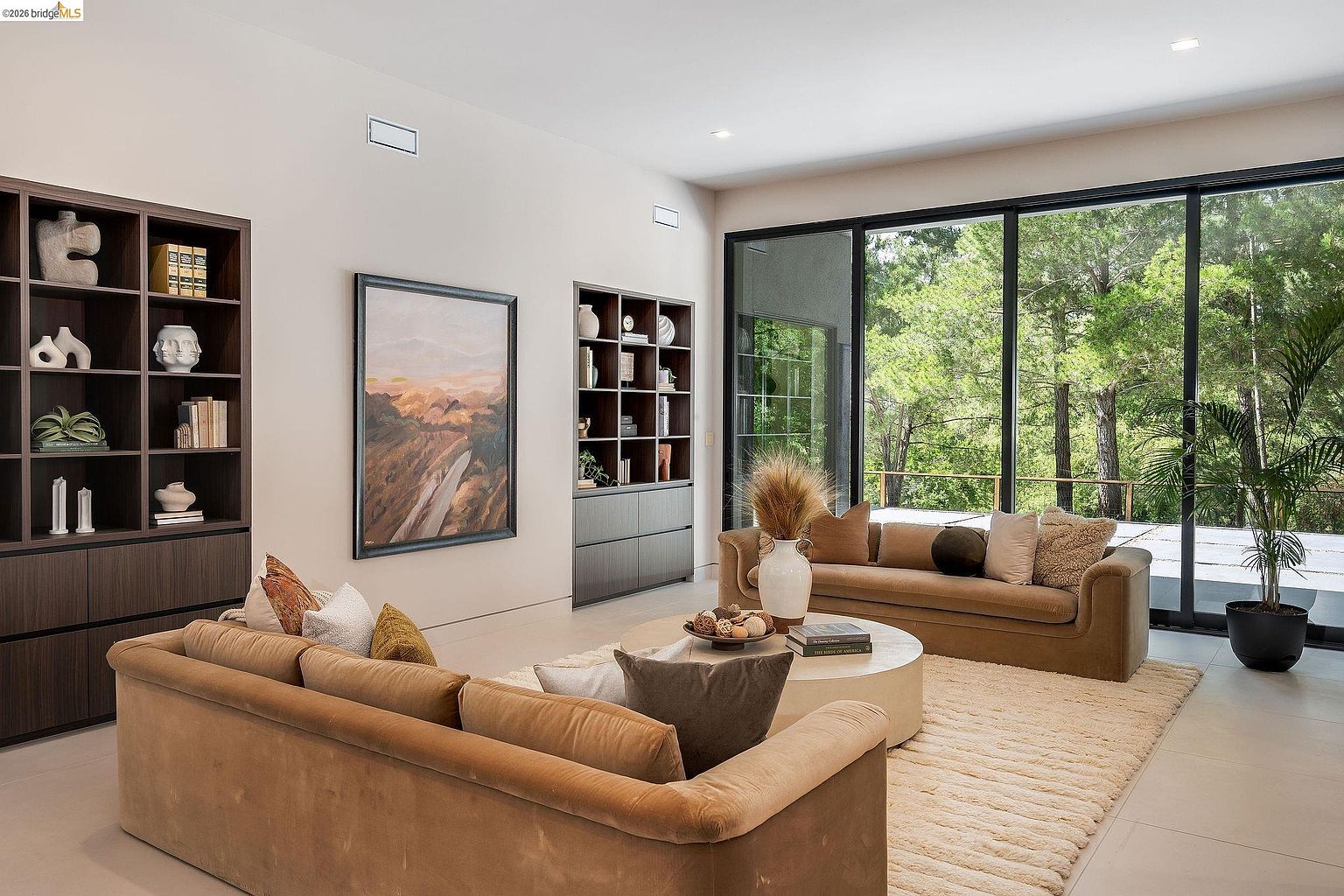 This sophisticated living room features a warm, neutral color palette with plush velvet sofas and a large circular coffee table centered on a textured area rug. The space is anchored by two floor-to-ceiling dark wood shelving units and a large abstract painting, while expansive glass sliding doors provide a seamless transition to the lush, tree-filled outdoor scenery. The overall aesthetic is modern, serene, and inviting, emphasizing natural light and high-end finishes.