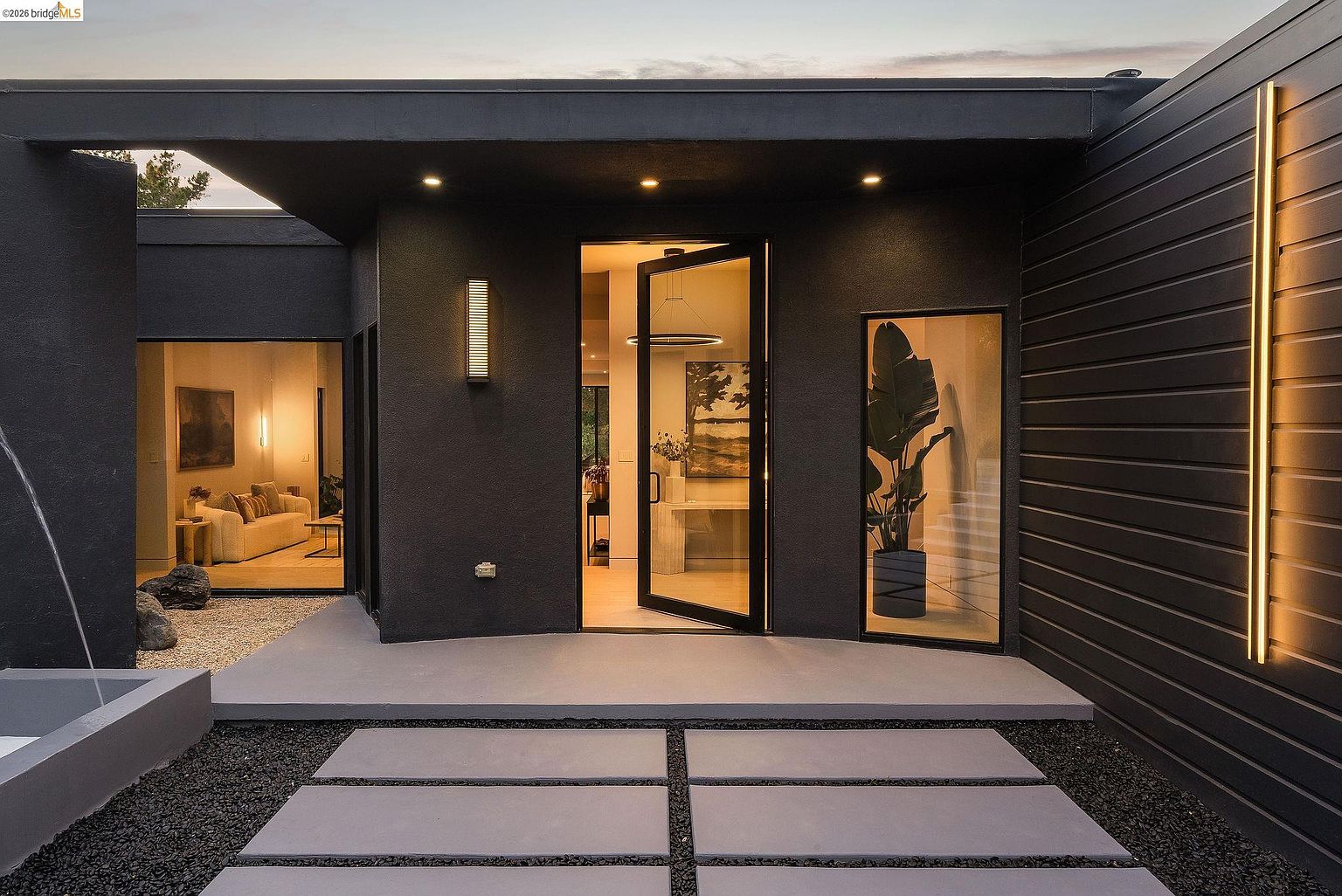 This striking modern entryway features a bold, dark charcoal facade contrasted by warm, vertical accent lighting and a sleek glass-paneled front door. The entrance is framed by large, rectangular concrete pavers set in dark gravel, leading to a covered porch that offers a glimpse into a sophisticated, well-lit interior. The composition emphasizes clean lines and a high-end, contemporary aesthetic, creating a welcoming yet dramatic first impression.