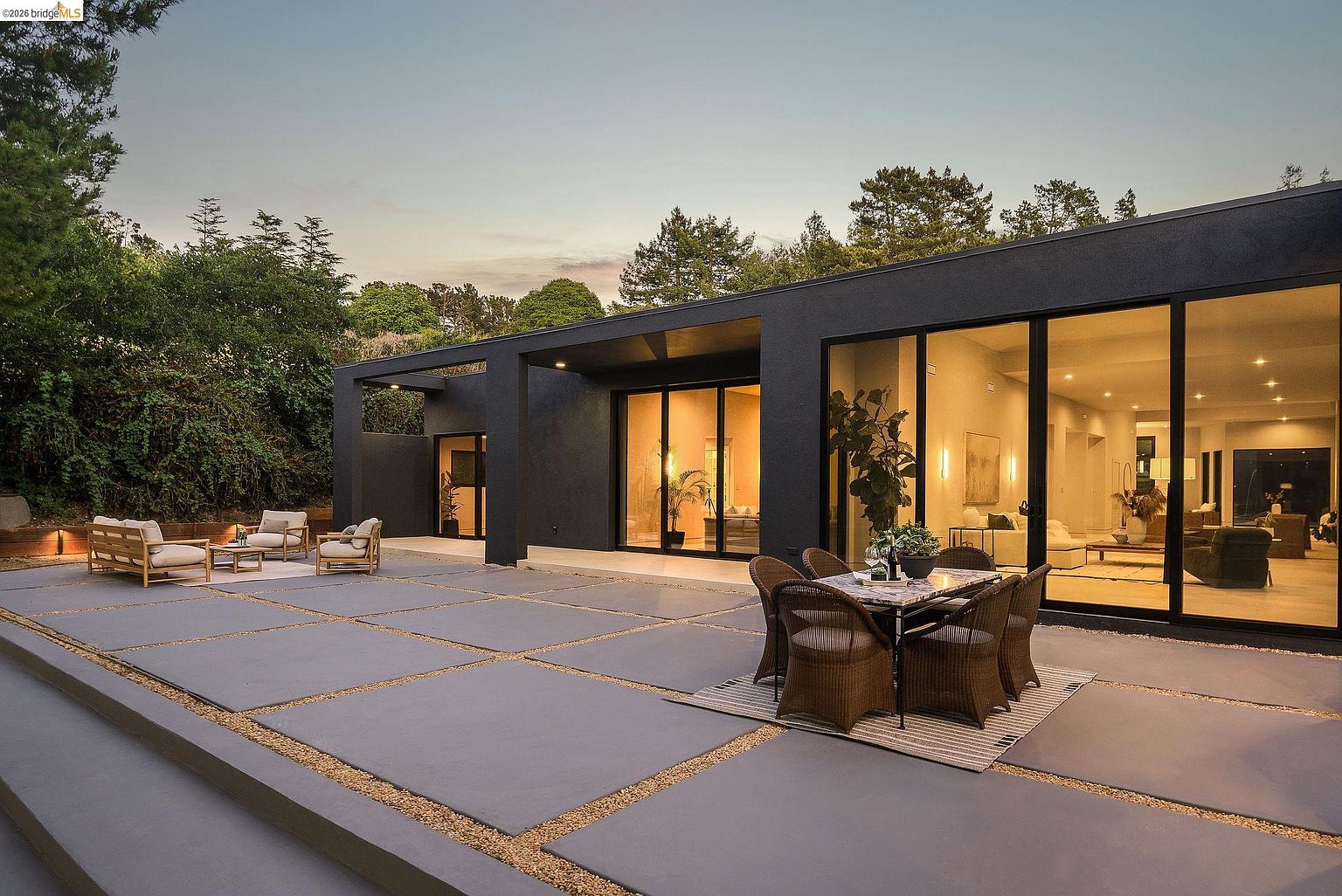 This modern, minimalist patio features large, square concrete pavers set in gravel, creating a clean and geometric outdoor living space. The area is furnished with a dining set and a separate lounge area, all seamlessly connected to the home's interior through expansive floor-to-ceiling glass sliding doors. The dark, sleek exterior of the house contrasts beautifully with the warm, inviting interior lighting, creating a sophisticated atmosphere perfect for evening entertaining.