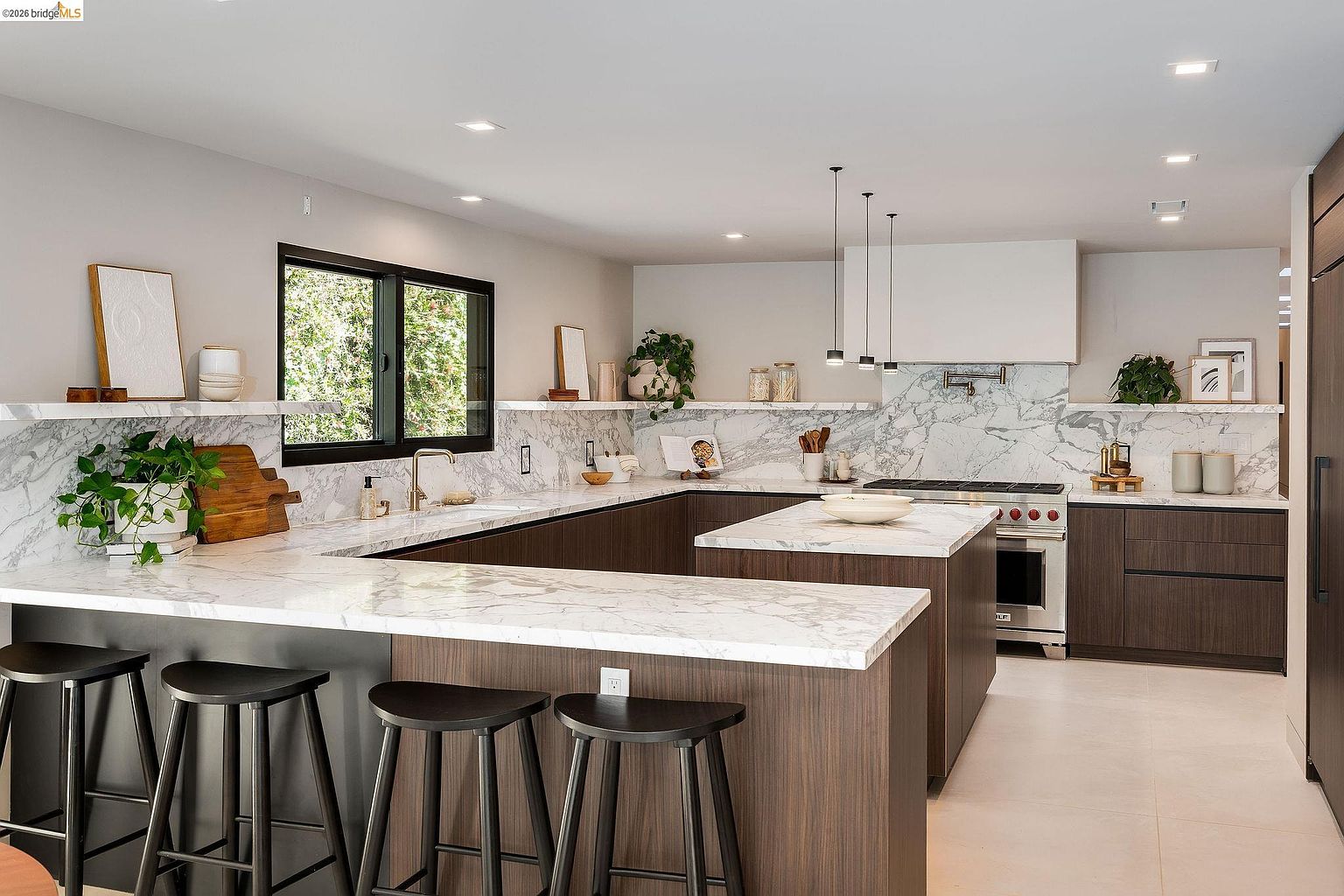 This modern, high-end kitchen features a spacious U-shaped layout with dark wood cabinetry and striking white marble countertops that extend into a large central island. The space is illuminated by sleek pendant lighting and recessed ceiling lights, highlighting the clean lines and contemporary aesthetic. A large window provides natural light, while the professional-grade range and integrated appliances emphasize the kitchen's functional elegance.