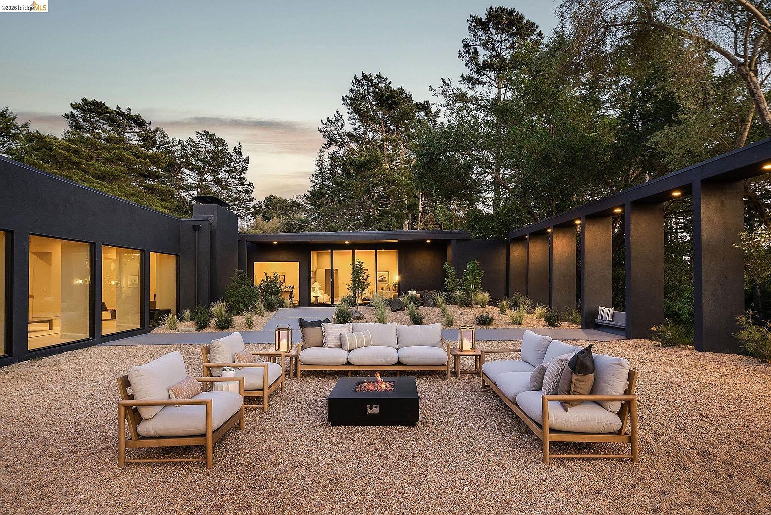 This stunning modern outdoor living space features a sleek, dark-painted architectural structure surrounding a gravel courtyard. A central fire pit is flanked by comfortable, light-toned lounge furniture, creating an inviting atmosphere for evening relaxation. The scene is framed by mature trees and warm, integrated lighting, emphasizing a sophisticated and tranquil contemporary aesthetic.