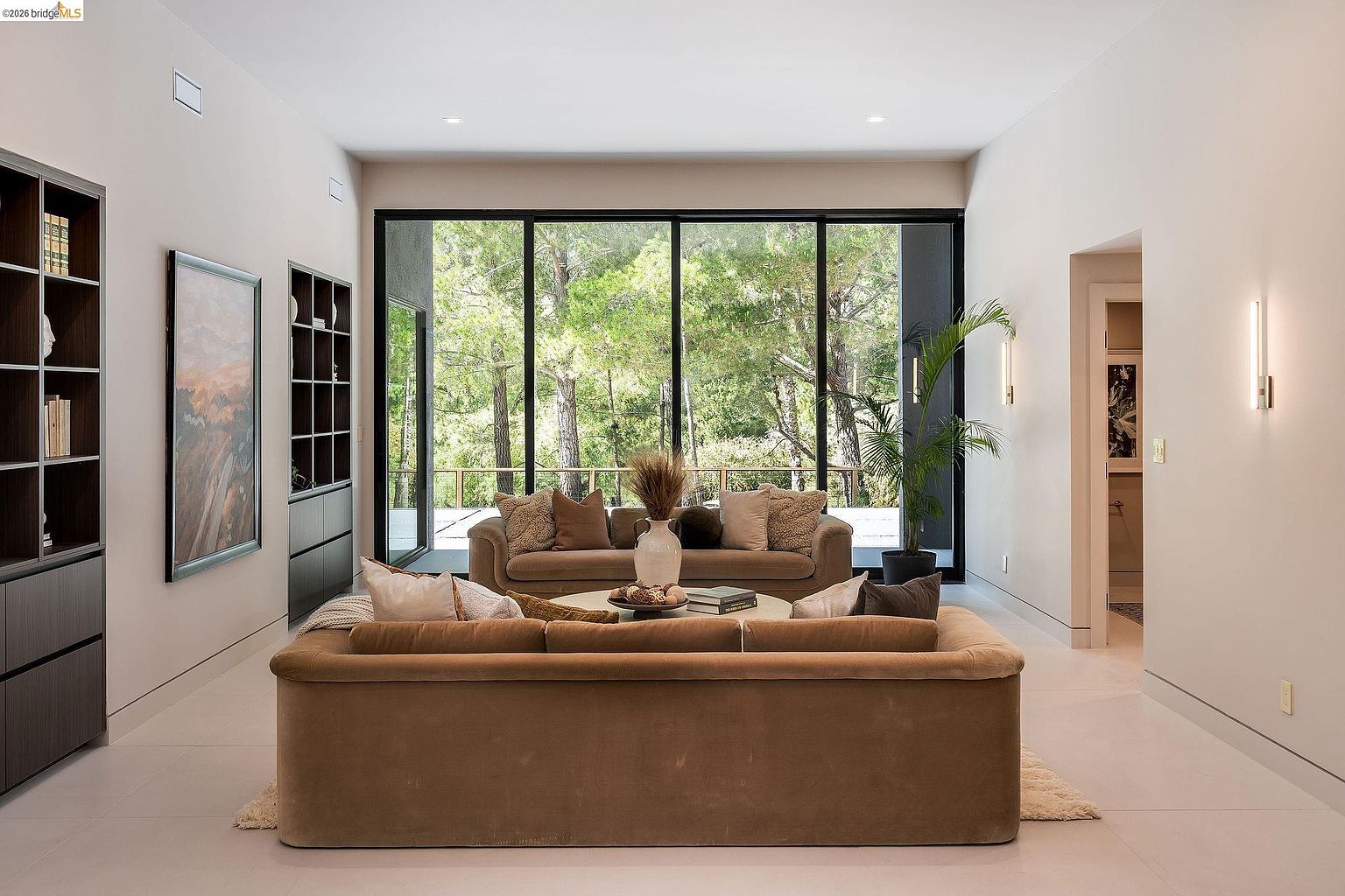 This sophisticated living room features a neutral, earthy color palette with plush, velvet-textured sofas and a central coffee table. A massive floor-to-ceiling glass wall provides a seamless connection to the lush, wooded exterior, flooding the space with natural light. The room is accented by built-in shelving, modern wall sconces, and a large piece of abstract art, creating a serene and upscale atmosphere.