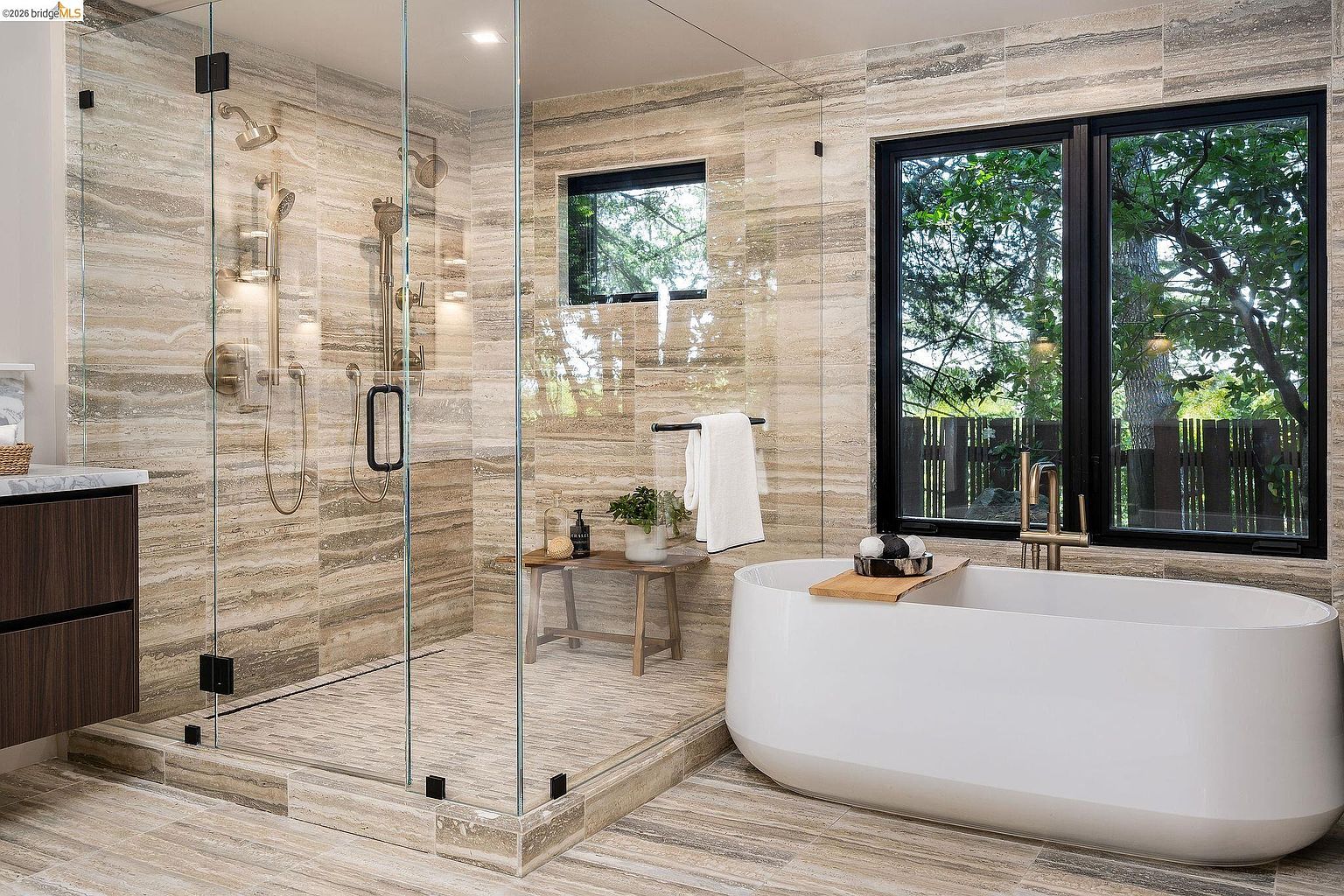 This luxurious primary bathroom features a seamless glass-enclosed walk-in shower with dual showerheads and a sleek, freestanding soaking tub positioned next to a large window. The space is finished with elegant, horizontal-veined stone tile throughout, creating a sophisticated and spa-like atmosphere. A small wooden bench adds a touch of organic warmth to the modern, high-end design.