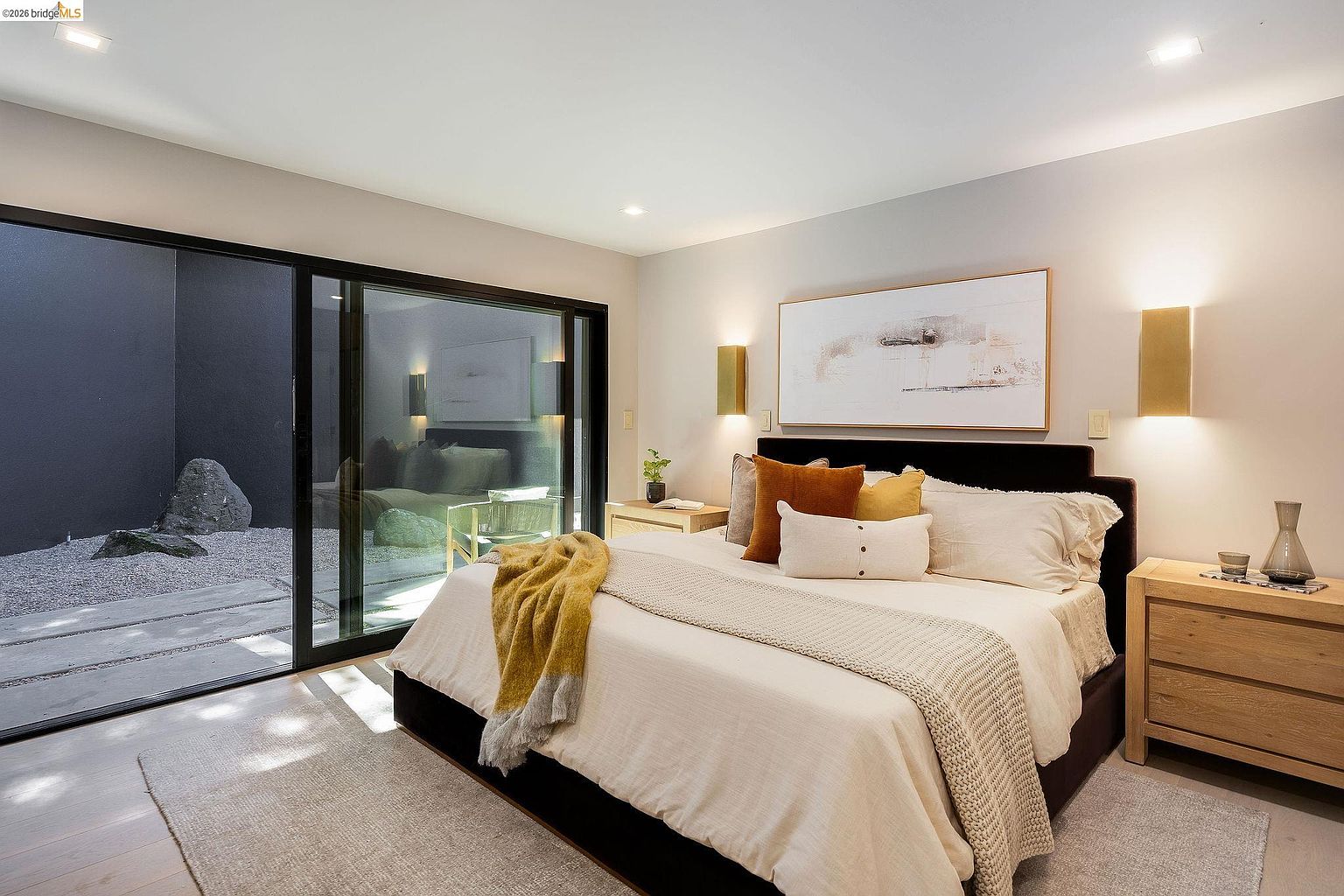 This elegant primary bedroom features a plush, dark-upholstered bed adorned with layered neutral and mustard-toned textiles, creating a sophisticated and inviting atmosphere. Large sliding glass doors open directly onto a serene, minimalist Zen-inspired courtyard, seamlessly blending indoor comfort with outdoor tranquility. The room is accented by warm wall sconces and a large abstract piece of art, emphasizing a modern, high-end aesthetic.