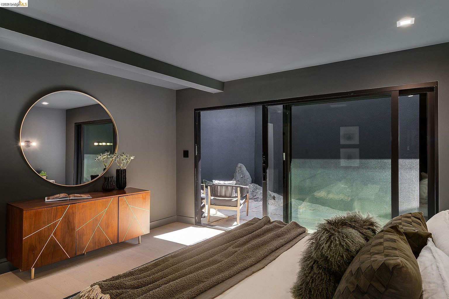 This sophisticated primary bedroom features a modern, moody aesthetic with dark grey walls and a warm wood-toned credenza topped with a large, circular mirror. A sliding glass door provides a seamless transition to a private, minimalist outdoor patio area, enhancing the room's sense of space and natural light. The foreground includes a plush bed with textured bedding, creating a cozy and inviting atmosphere perfect for a high-end residential listing.