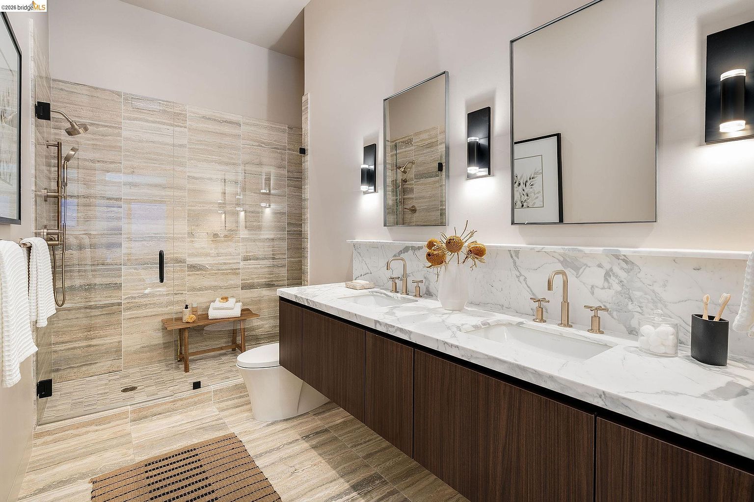 This luxurious primary bathroom features a sleek, modern design with a floating dark wood vanity topped by a white marble countertop and dual sinks. A spacious walk-in shower with textured stone-look tiles and a glass enclosure serves as a focal point, complemented by minimalist wall sconces and rectangular mirrors. The overall aesthetic is sophisticated and spa-like, emphasizing clean lines and high-end finishes.
