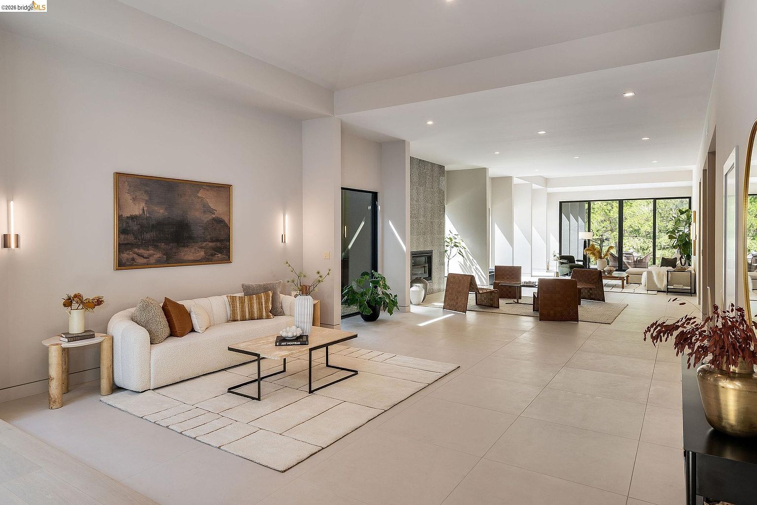 This expansive, open-concept living area showcases a sophisticated modern aesthetic with neutral tones and high ceilings. The space features a plush cream sofa, a geometric coffee table, and a large area rug, leading the eye toward a secondary seating area and floor-to-ceiling windows that invite natural light. The minimalist design is complemented by a textured stone fireplace and elegant, understated decor, creating a serene and luxurious atmosphere.