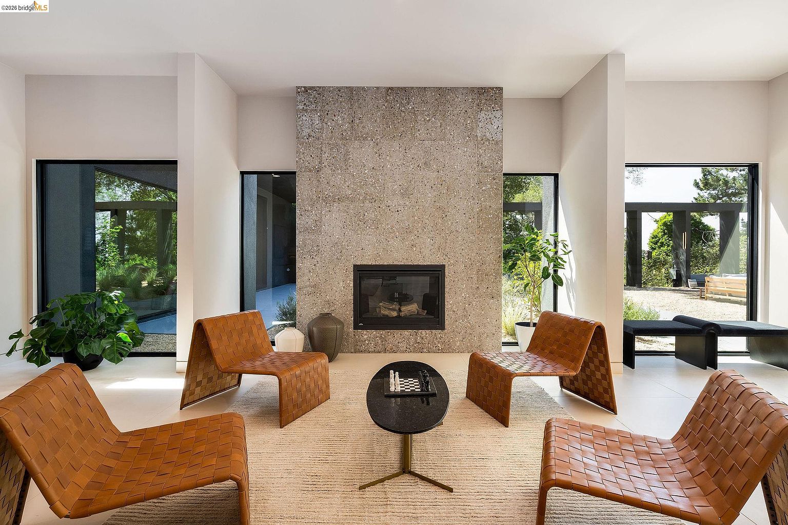 This sophisticated living room features a striking floor-to-ceiling stone fireplace as its central focal point, flanked by large windows that invite abundant natural light and outdoor views. The space is furnished with four modern, woven leather lounge chairs arranged around a sleek, circular black coffee table on a textured area rug. The minimalist design, neutral color palette, and open layout create an atmosphere of refined, contemporary elegance.
