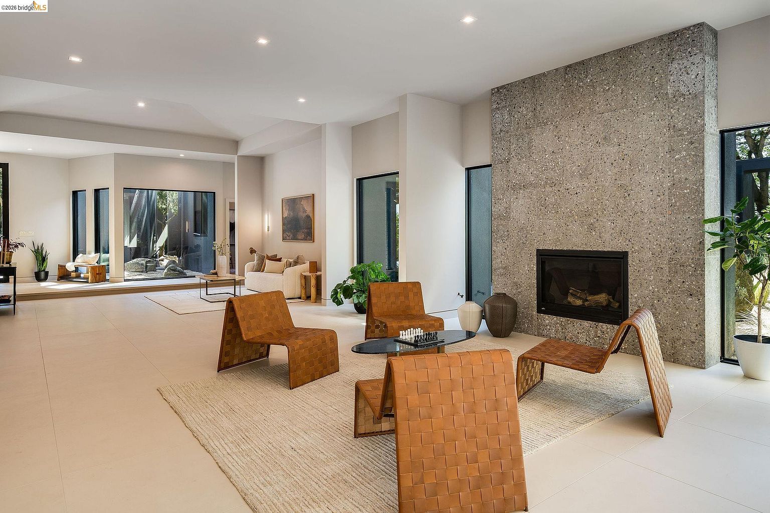 This spacious, modern living room features a striking floor-to-ceiling stone fireplace as its focal point, complemented by minimalist, light-colored tile flooring. The room is furnished with unique, woven leather lounge chairs arranged around a sleek glass coffee table, creating an inviting and sophisticated atmosphere. Large windows and an open-concept layout allow for seamless transitions to an adjacent seating area, emphasizing a bright, airy, and high-end architectural design.