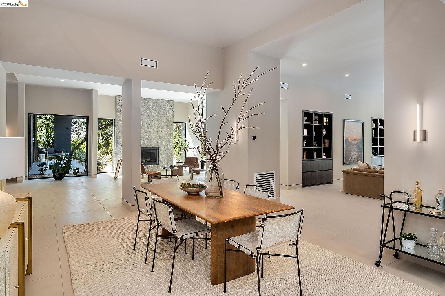 This elegant dining area features a sleek wooden table surrounded by modern, minimalist chairs, set atop a textured area rug. The open-concept design flows seamlessly into a bright living space with a stone fireplace and large glass doors that invite natural light and views of the outdoors. The neutral color palette and clean lines create a sophisticated, airy atmosphere perfect for contemporary living.
