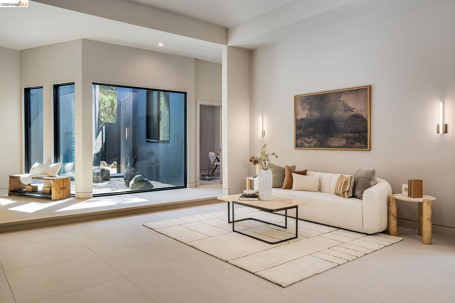 This sophisticated living room features a minimalist, contemporary design with a neutral color palette and high ceilings. A plush, curved sofa sits atop a textured area rug, complemented by a marble-topped coffee table and a large, moody landscape painting on the wall. The space is anchored by a striking floor-to-ceiling window that frames a serene, rock-filled courtyard, creating a seamless connection between the interior and exterior.