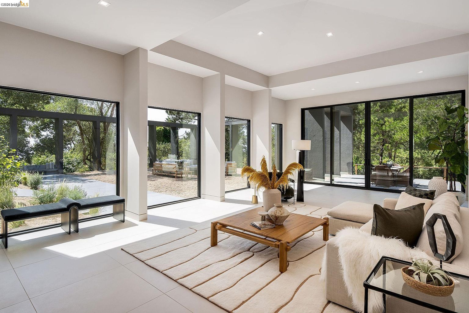 This bright and airy living room features a minimalist, contemporary design with expansive floor-to-ceiling windows that seamlessly blend the indoor space with the lush outdoor greenery. The room is anchored by a neutral-toned sofa, a textured area rug, and a warm wooden coffee table, creating a sophisticated yet inviting atmosphere. The open-concept layout and high ceilings emphasize the home's spaciousness and connection to nature.
