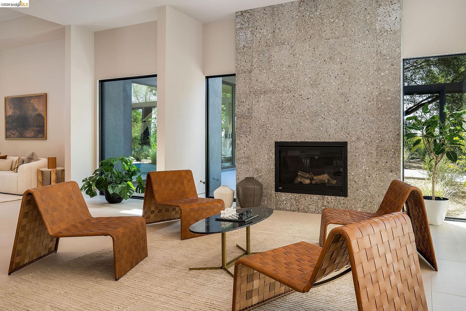This sophisticated living room features a striking floor-to-ceiling textured stone fireplace as its central focal point. The space is furnished with four modern, woven leather lounge chairs arranged around an elegant oval coffee table, all set upon a neutral-toned area rug. Large floor-to-ceiling windows provide abundant natural light and seamless views of the outdoor landscape, creating an airy and contemporary atmosphere.