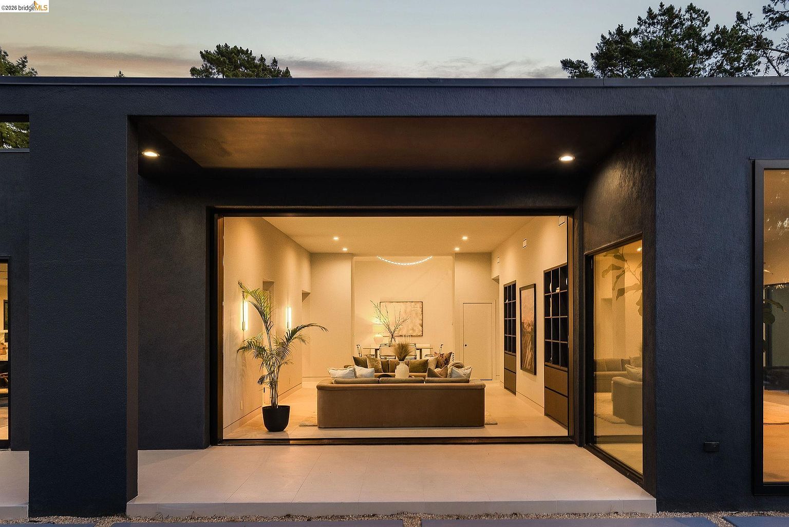 This image captures a sleek, modern patio transition leading into a warm, inviting living space through expansive sliding glass doors. The dark, minimalist exterior facade contrasts beautifully with the soft, ambient interior lighting, creating a sophisticated and cinematic atmosphere. The scene highlights a seamless indoor-outdoor flow, perfect for evening entertaining, with a clear view of the tastefully decorated living area beyond.