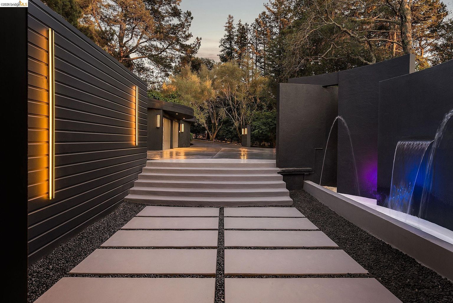 This striking modern entryway features a dramatic, dark-toned architectural design with vertical accent lighting and a cascading water feature illuminated by purple and blue LED lights. A set of wide, clean-lined concrete steps leads up to a paved driveway area, framed by dark horizontal siding and lush, mature trees in the background. The composition offers a sophisticated, high-end aesthetic that emphasizes luxury and contemporary landscape design.