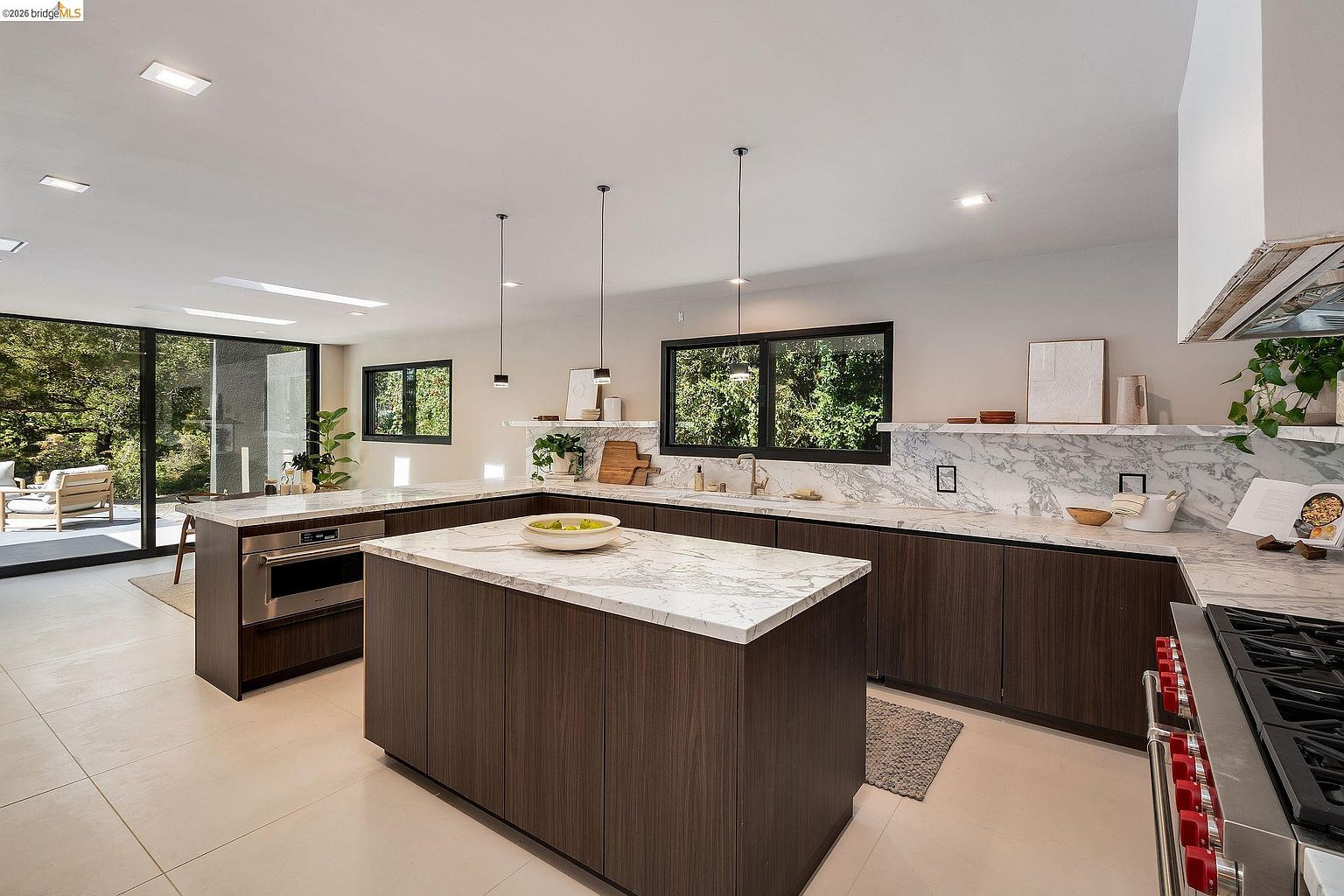 This modern, open-concept kitchen features dark wood cabinetry contrasted by elegant white marble countertops and a matching backsplash. A large central island serves as a focal point, complemented by sleek pendant lighting and high-end stainless steel appliances. The space is bright and airy, with floor-to-ceiling glass doors that seamlessly connect the interior to the outdoor patio area.