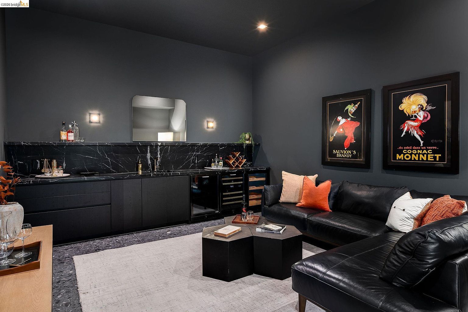 This sophisticated home bar and lounge area features a sleek, dark aesthetic with black marble countertops, matte black cabinetry, and a matching leather sofa. The space is accented by two vintage-style framed posters, a geometric coffee table, and warm wall sconce lighting, creating an intimate and luxurious atmosphere. The perspective is a wide, eye-level shot that captures the seamless blend of functional bar space and comfortable seating.