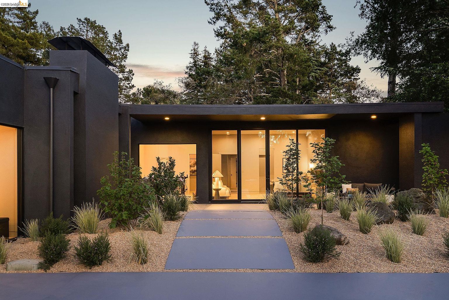 This striking modern home features a bold, dark charcoal exterior with clean lines and a minimalist aesthetic. A wide, paved walkway leads directly to a large glass-paneled entryway, flanked by thoughtfully landscaped desert-style gardens with ornamental grasses and small trees. The warm interior lighting glowing through the glass creates an inviting contrast against the dramatic, moody facade.