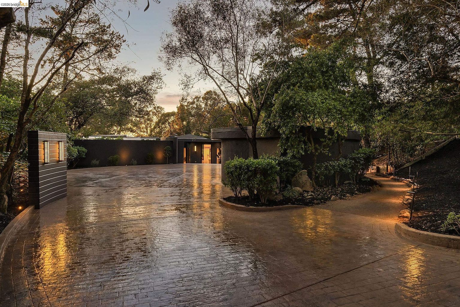 This striking entryway features a wide, wet-look stamped concrete driveway leading toward a modern, minimalist home entrance framed by dark, textured walls and lush, mature landscaping. The scene is captured at dusk, with warm, recessed lighting casting dramatic shadows and highlighting the sleek architectural lines of the property. The perspective is low and wide, emphasizing the grand, private approach to the residence and creating an inviting, sophisticated atmosphere.
