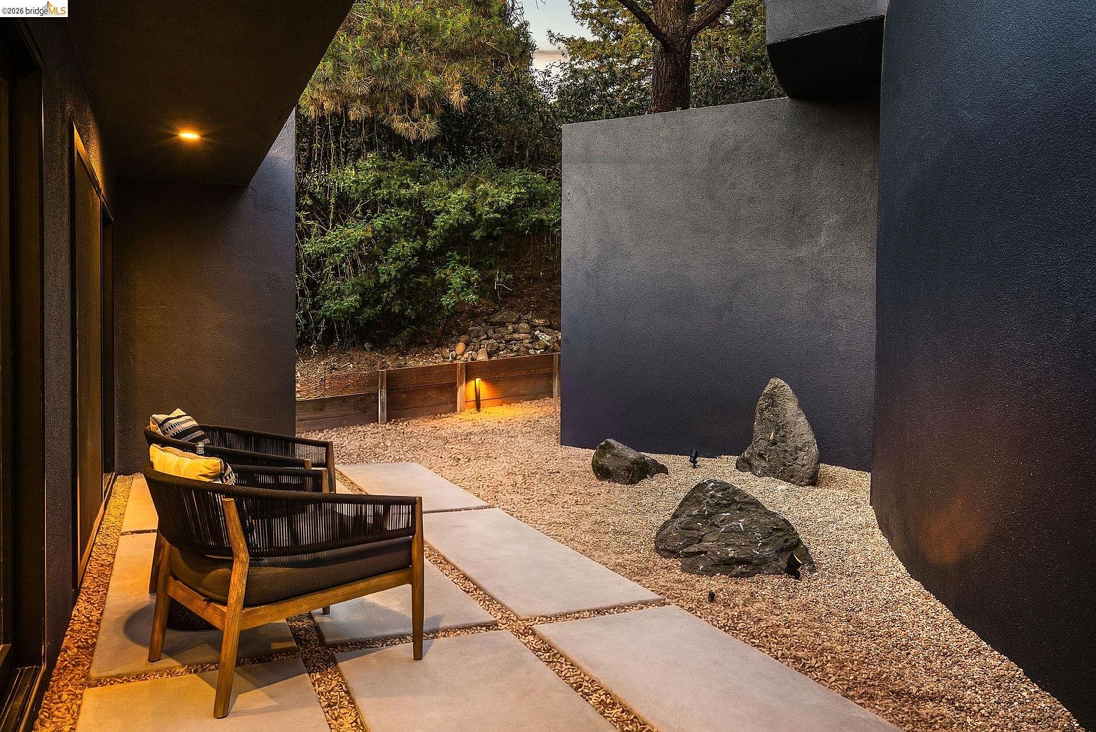 This modern, minimalist patio features a sleek design with dark, charcoal-colored walls and large concrete pavers set into a gravel bed. Two contemporary armchairs with dark woven frames and cushions provide a comfortable seating area, while a small rock garden adds a touch of Zen-inspired landscaping. The perspective is from under a covered overhang, looking out toward the serene, private outdoor space.