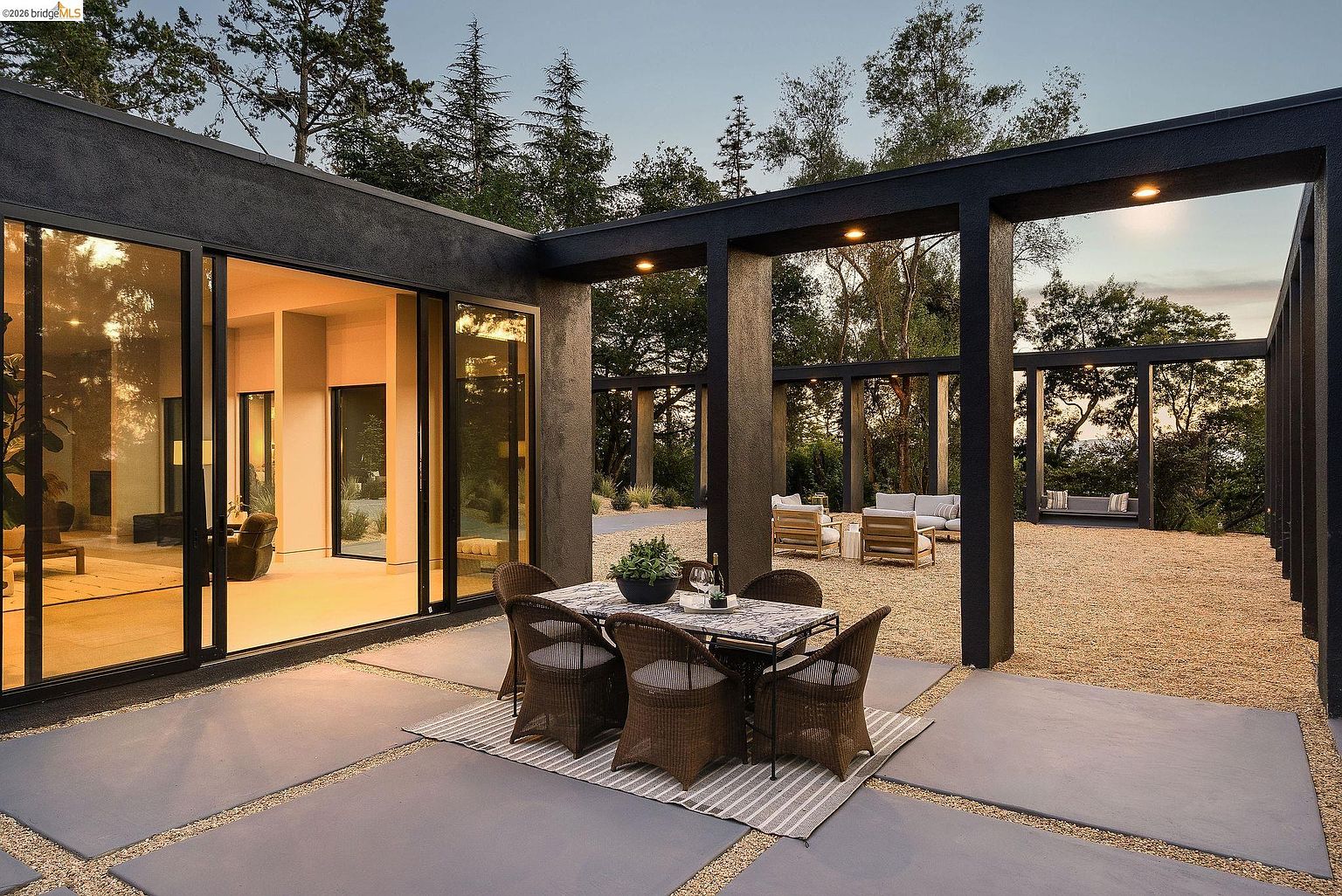 This sophisticated outdoor patio features a modern, minimalist design with large concrete pavers and a gravel courtyard framed by a striking black architectural pergola. A dining set with wicker chairs sits on a striped rug in the foreground, while a comfortable lounge area is visible in the background under the open-air structure. The seamless transition between the indoor living space and the outdoor area, combined with the warm evening lighting, creates an inviting and luxurious atmosphere.