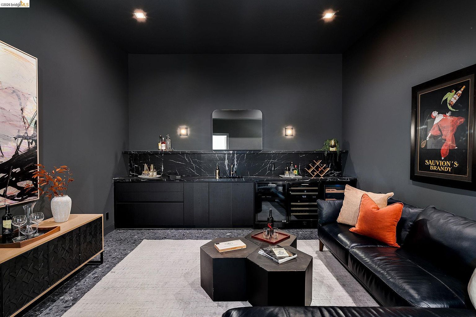 This sophisticated lounge area features a moody, monochromatic aesthetic with dark charcoal walls, black marble countertops, and sleek cabinetry. The space is anchored by a plush black leather sofa accented with vibrant orange pillows, a pair of geometric coffee tables, and a large, light-colored area rug that provides a striking contrast. A framed vintage 'Sauvion's Brandy' poster and abstract wall art add character to this elegant, high-end entertainment space.