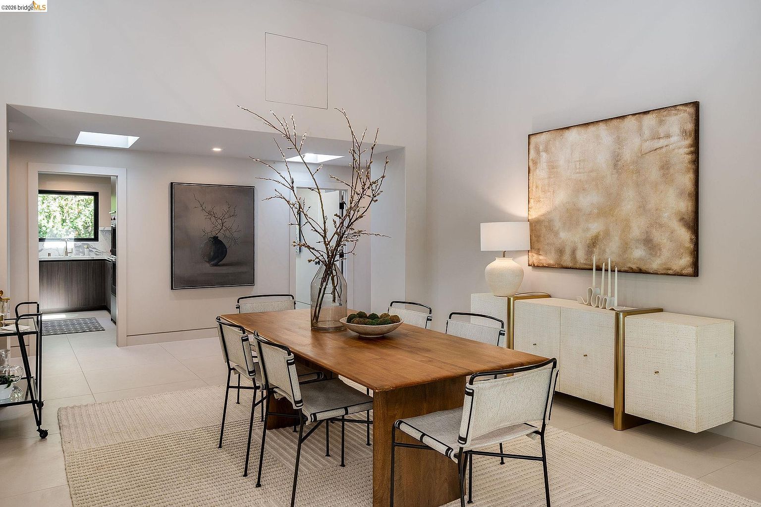 This elegant dining room features a warm, natural wood table surrounded by modern chairs with a minimalist aesthetic, set atop a textured area rug. A sleek, cream-colored sideboard with gold accents sits against the wall, topped with a contemporary lamp and abstract artwork, while a large vase with branches serves as a striking centerpiece. The space is bright and airy, with high ceilings and a clear view into the adjacent kitchen, creating a sophisticated and inviting atmosphere.