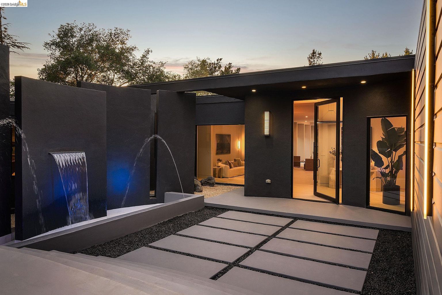 This striking modern entryway features a dramatic dark facade with a cascading water feature and a minimalist concrete paver walkway. The open glass door invites viewers into a warm, well-lit interior, creating a sophisticated contrast between the moody exterior and the inviting home. The architectural design emphasizes clean lines and high-end materials, offering a luxurious and welcoming first impression.