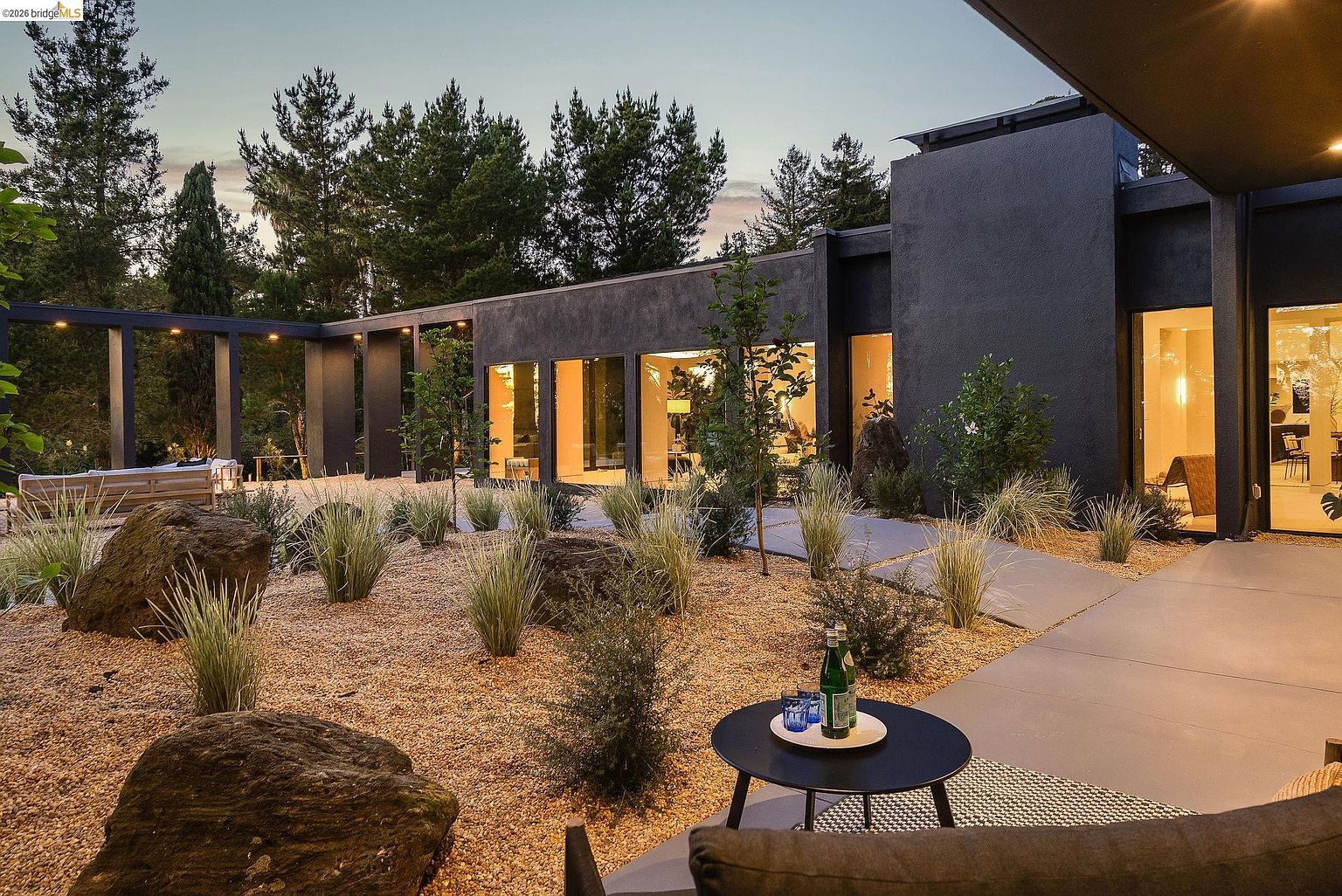 This image captures a sophisticated modern patio area featuring a sleek, dark-toned architectural structure that frames a minimalist xeriscaped garden. The foreground showcases a small, contemporary round table with refreshments, set against a backdrop of gravel landscaping, large decorative boulders, and floor-to-ceiling glass walls that reveal a warm, inviting interior. The perspective is low and intimate, emphasizing the seamless transition between the refined outdoor living space and the elegant, illuminated home interior.