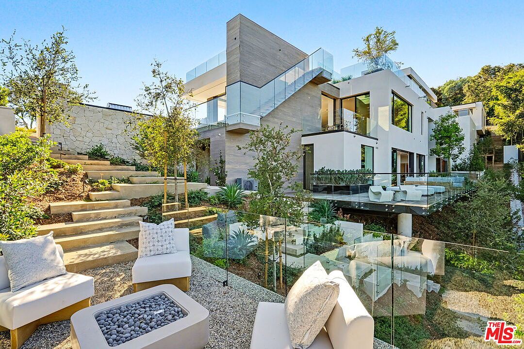 This contemporary multi-level residence features a sophisticated architectural design with clean lines, light-toned stucco, and textured wood-look siding. The rear exterior showcases expansive glass railings, a cantilevered deck, and a tiered landscape with concrete steps leading down to a gravel patio area equipped with a modern fire pit. The composition captures a seamless transition between indoor living spaces and the terraced outdoor environment, emphasizing a luxurious, modern lifestyle.