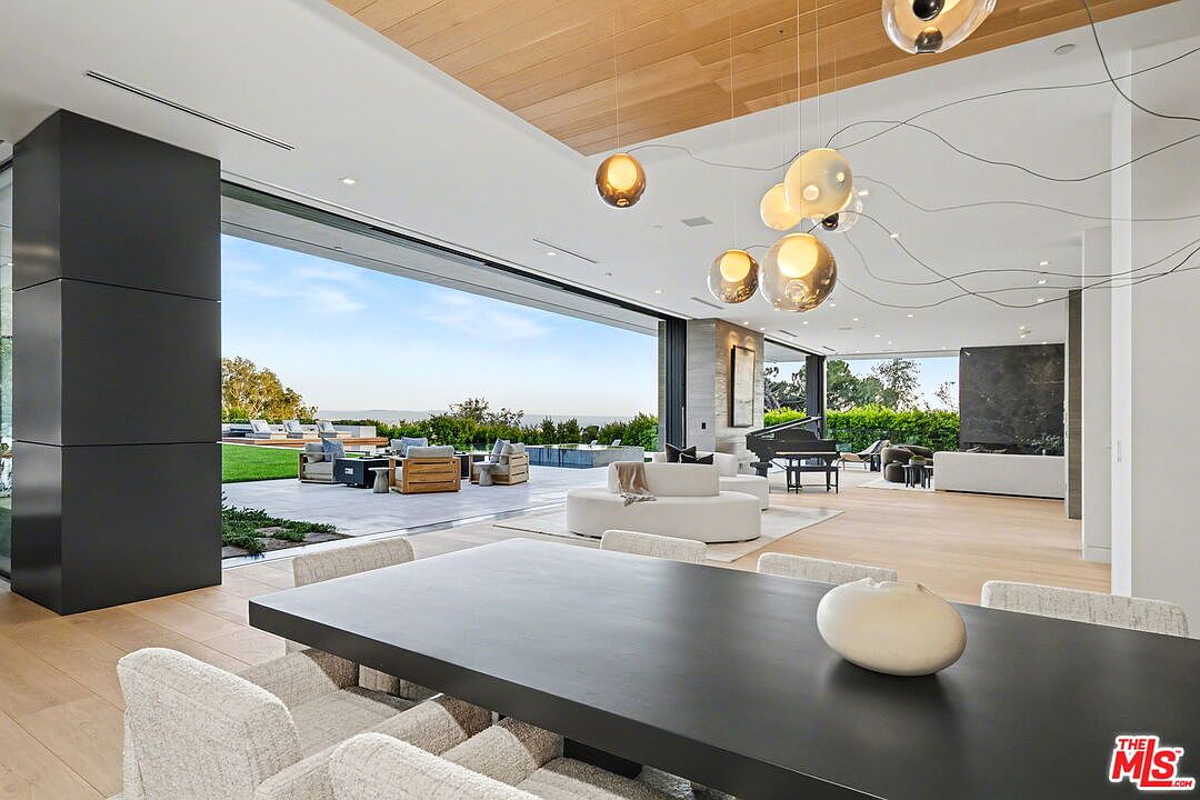 This expansive, open-concept living space features a dining area in the foreground with a large dark table and light-toned chairs. The room flows seamlessly into a bright living area with a curved sofa and a grand piano, all illuminated by a unique cluster of spherical pendant lights. Massive glass walls slide open to reveal a spacious outdoor patio and a scenic landscape, creating a fluid transition between indoor and outdoor living.