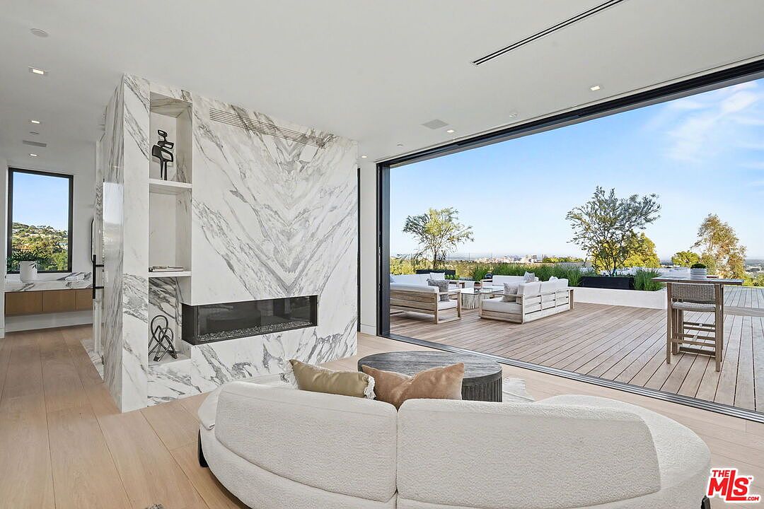This modern living room features a curved, light-colored sofa positioned before a striking stone-clad chimney breast with a built-in linear fireplace and integrated shelving. Large, sliding glass doors open fully to a spacious wooden deck, creating a seamless transition between the indoor living area and the outdoor terrace. The room is finished with light wood flooring and offers an expansive, elevated view of the surrounding landscape.