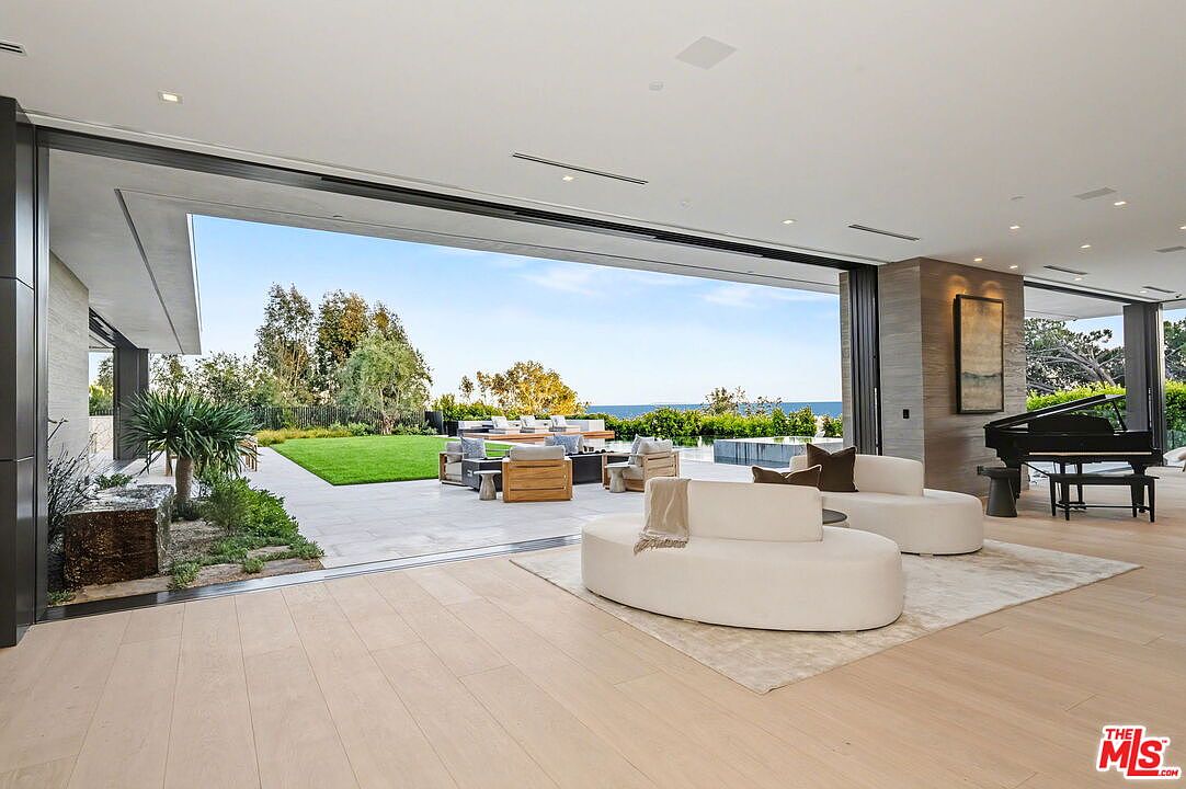 This expansive, modern living room features an open-concept design that seamlessly transitions to an outdoor patio through massive, retractable glass walls. The interior is furnished with curved, contemporary seating on a neutral area rug, while a grand piano sits to the right, adding a touch of elegance to the space. The outdoor area includes a manicured lawn, comfortable lounge furniture, and a scenic view of the ocean, creating a sophisticated indoor-outdoor living experience.