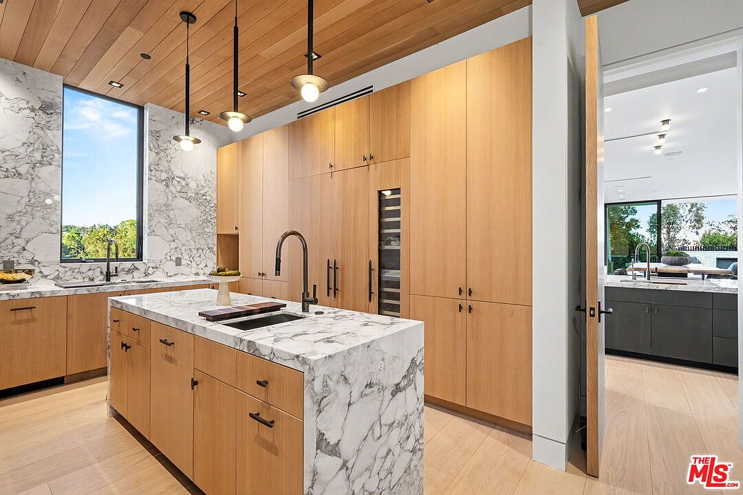 This modern kitchen features a large central island with a stone countertop and integrated sink, complemented by light wood cabinetry and a matching wood-paneled ceiling. A tall window with a stone surround provides natural light, while three pendant lights hang above the island. The perspective captures the main kitchen area with a glimpse into an adjacent room through an open doorway, showcasing a cohesive and high-end design.