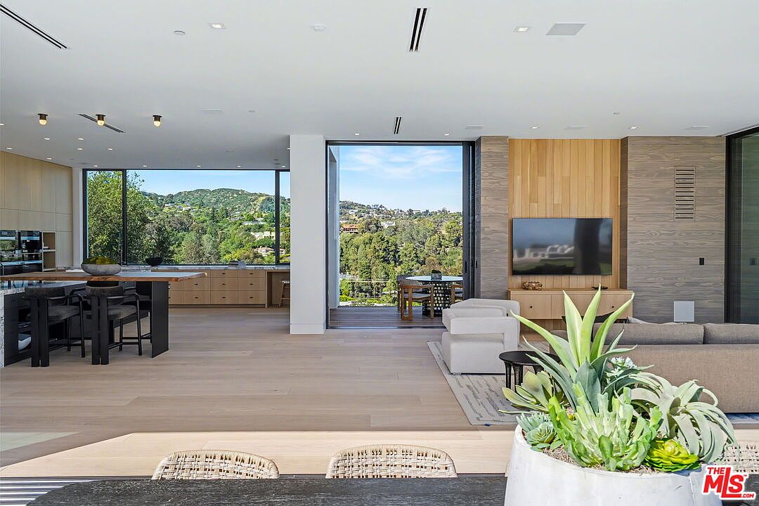 This expansive open-concept living space features light wood flooring and a seamless transition to an outdoor deck through large sliding glass doors. The room includes a comfortable seating area with a television mounted on a wood-paneled wall, adjacent to a modern kitchen with a large island. The perspective is from a slightly elevated position, looking across the living area toward the scenic hillside view visible through the glass.