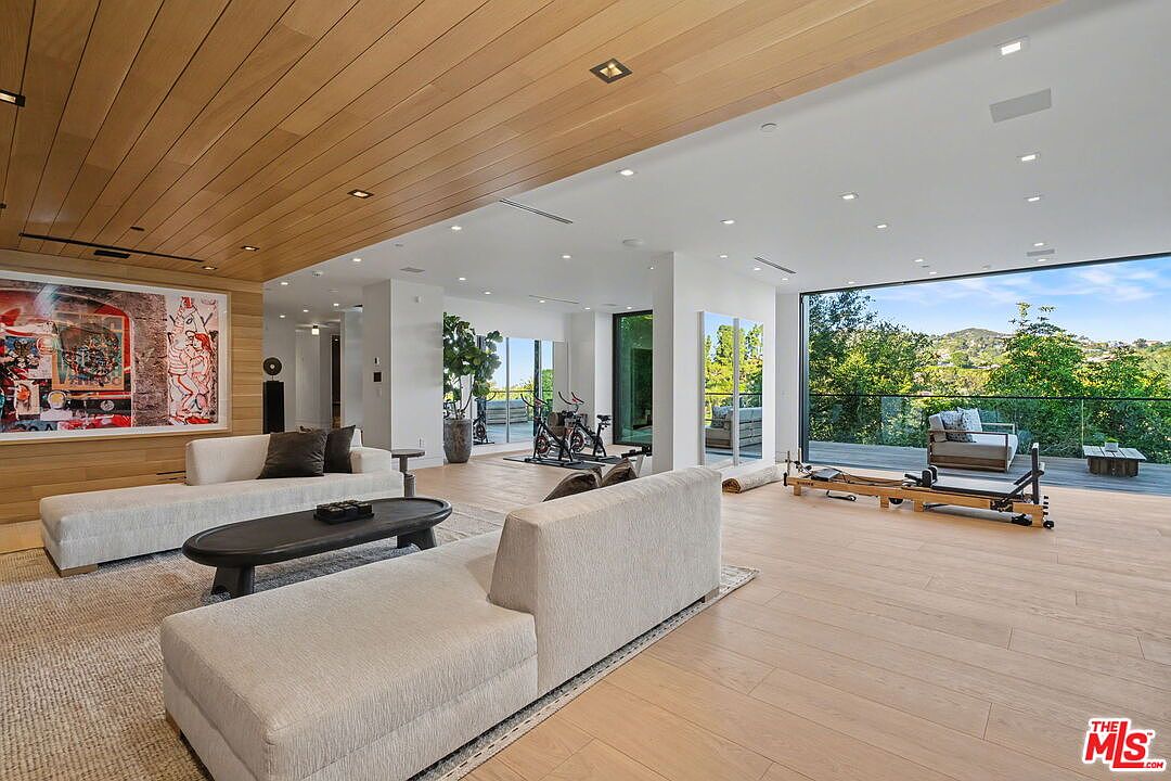 This expansive, open-concept living area features light wood flooring and a matching wood-paneled ceiling, creating a warm and cohesive aesthetic. The space is furnished with a large, neutral-toned sectional sofa and a dark oval coffee table, while a dedicated fitness area with exercise bikes and a Pilates reformer is integrated into the background. Large glass sliding doors open onto an outdoor deck, providing a seamless transition to the exterior and offering panoramic views of the surrounding hills.