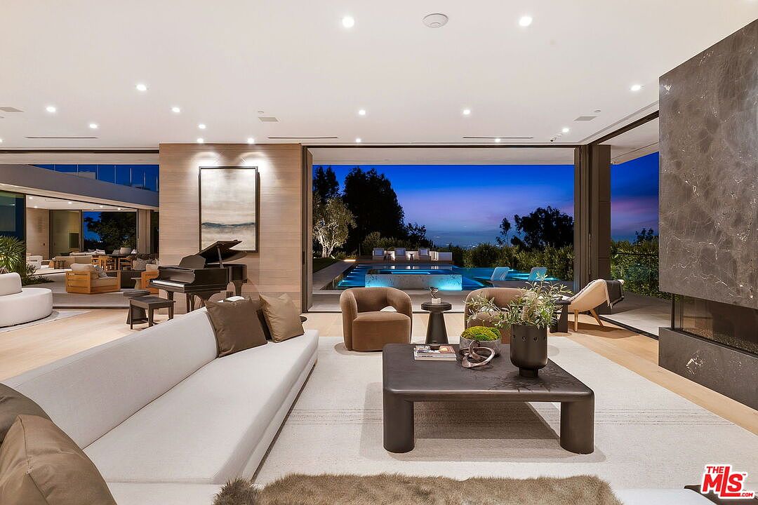 This expansive, modern living room features a neutral color palette and an open-concept design that seamlessly transitions to an outdoor pool area through large glass sliding doors. The space is furnished with a long, low-profile sofa, a dark coffee table, and two plush armchairs, with a grand piano positioned near a wood-paneled wall. A prominent stone-clad chimney breast anchors the right side of the room, creating a sophisticated and airy atmosphere perfect for luxury entertaining.