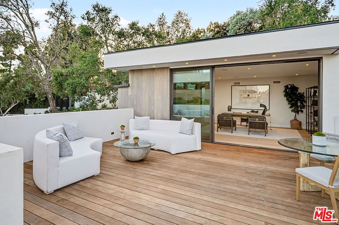 This contemporary outdoor deck features warm-toned wood plank flooring and a minimalist white perimeter wall, creating a seamless transition to the interior office space through large sliding glass doors. The area is furnished with modern white curved seating and a central metallic coffee table, while a glass-topped dining table sits to the right. The scene is framed by lush green trees in the background, offering a serene and sophisticated atmosphere for outdoor relaxation or professional work.