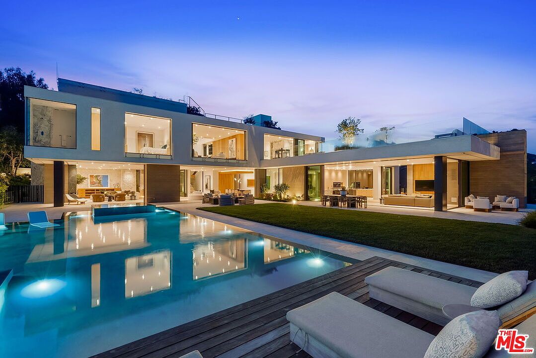 This contemporary luxury residence features a sprawling, multi-level architectural design with extensive glass walls that seamlessly connect the indoor living spaces to the outdoor patio. The property includes a large, rectangular swimming pool with integrated lounge areas, set against a manicured lawn and a wooden deck with modern sun loungers. The twilight setting highlights the warm interior lighting, creating an inviting and sophisticated atmosphere for evening entertaining.
