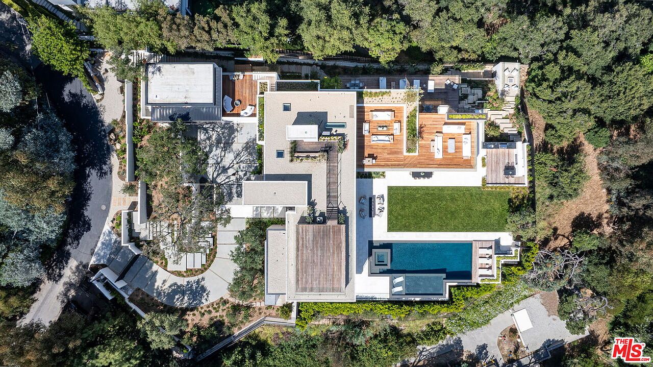 This high-angle aerial perspective captures a sprawling, modern luxury estate nestled within a lush, wooded landscape. The property features a sophisticated layout with multiple interconnected flat-roof structures, expansive wooden decking, a rectangular swimming pool, and a manicured lawn. The design emphasizes seamless indoor-outdoor living, with various outdoor seating areas and pathways integrated into the natural terrain.