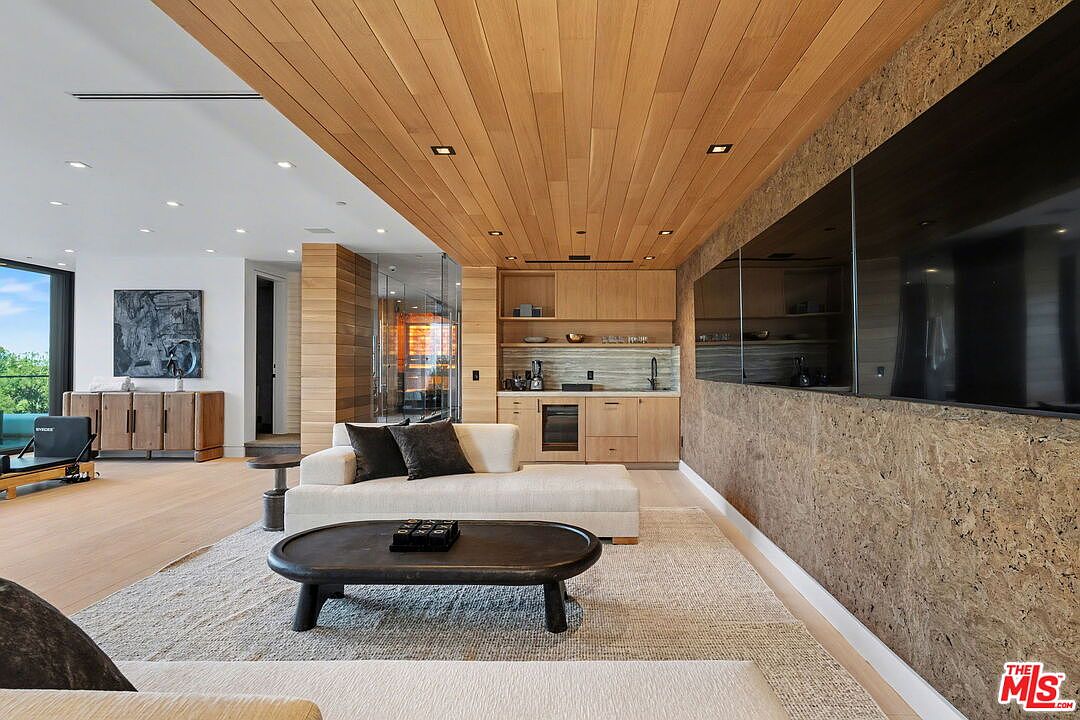 This contemporary living space features a warm, light wood-paneled ceiling and a textured, organic-toned accent wall that houses a large, integrated television. A sleek, light-colored sectional sofa sits atop a neutral area rug, centered around a dark, oval-shaped coffee table. In the background, a built-in wet bar with light wood cabinetry and a wine refrigerator adds functionality, while the open-concept layout flows seamlessly into an adjacent area with a sideboard and exercise equipment.