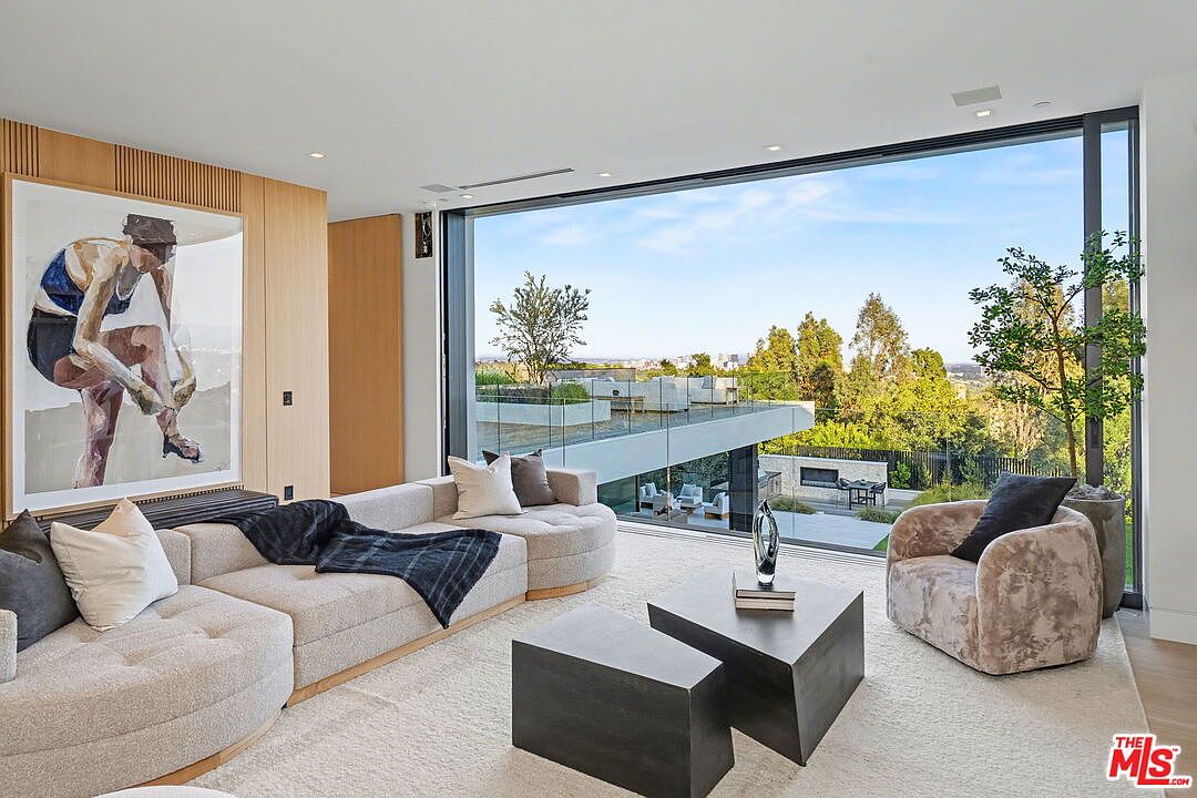 This sophisticated living room features a large, neutral-toned sectional sofa and a textured armchair arranged around two dark, geometric coffee tables. A prominent piece of figurative art hangs on the wood-paneled wall, while expansive floor-to-ceiling glass walls provide an unobstructed view of the outdoor terrace and surrounding landscape. The space exudes a modern, minimalist aesthetic with a focus on clean lines and a seamless connection to the exterior environment.