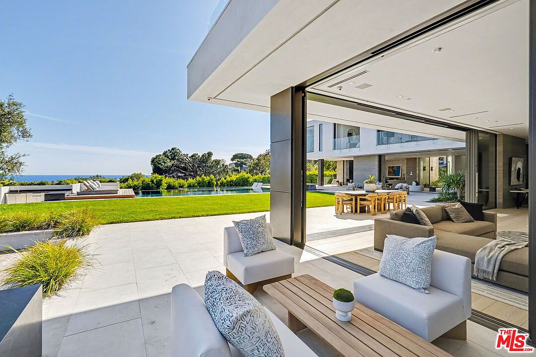 This luxurious indoor-outdoor living space features a seamless transition between a modern interior lounge and a sprawling stone patio. The area is furnished with contemporary white seating and a light wood coffee table, overlooking a manicured lawn, a sleek infinity-edge pool, and a distant ocean view. Large, retractable glass walls open the home to the elements, creating an expansive and airy atmosphere perfect for high-end entertaining.
