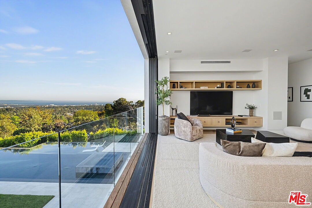 This sophisticated living room features an open-concept design that seamlessly transitions to an outdoor pool area through expansive, retractable glass walls. The interior is furnished with a curved, light-toned sofa and a contemporary armchair, complemented by a custom light wood media console and shelving unit. The space offers a panoramic view of the surrounding landscape and distant coastline, creating a bright and airy atmosphere.
