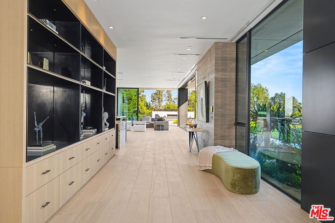 This modern hallway features light wood flooring and a long, built-in storage unit with dark shelving and light wood cabinetry. To the right, large windows provide an expansive view of the outdoor landscape, while a soft, curved bench sits beneath the glass. The perspective looks down the corridor toward an open-concept living area, creating a bright and airy transition space.