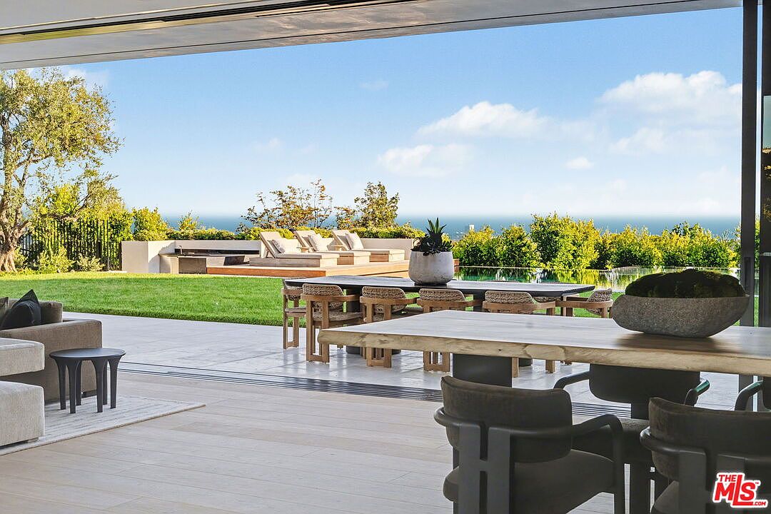 This indoor-outdoor living space features a seamless transition from a covered patio to a lush backyard with a swimming pool and ocean views. The foreground includes a wooden dining table and chairs, while the patio area is furnished with a comfortable sofa and a small side table. The open-concept design emphasizes a bright, airy atmosphere, perfect for entertaining and enjoying the coastal scenery.