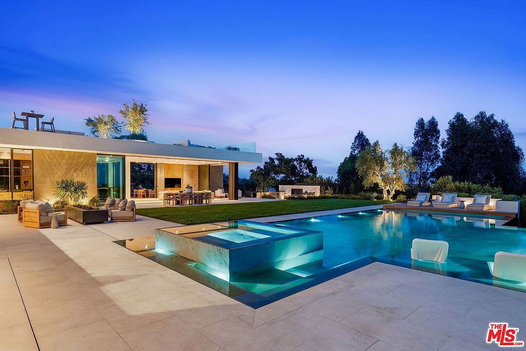 This stunning contemporary residence features a seamless indoor-outdoor transition with expansive glass walls opening onto a manicured lawn and a sophisticated infinity-edge pool. The backyard is anchored by a prominent, elevated spa that glows with integrated lighting, complemented by a spacious stone patio and lounge seating. The scene is captured at twilight, highlighting the architectural lighting and the serene, high-end atmosphere of the property.